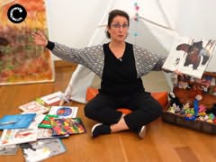 Senhora de braços abertos segurando um livro sentada de pernas cruzadas com mala com bonecos e livros ao lado