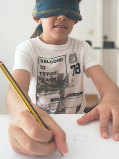 Rapaz vendado sentado a mesa com folha de papel e com lápis na mão