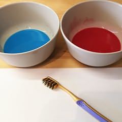 Uma tigela com tinta azul e outra com vermelha e uma folha de papel com uma escova de dentes por cima