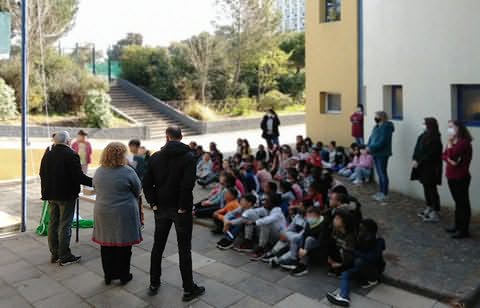 Muitas crianças sentadas no chão de pátio de escola com alguns adultos de pé