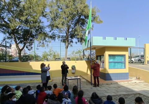 Bandeira verde no topo de haste com adulto puxando respetiva corda, dois adultos aplaudindo e muitas crianças sentadas no chão