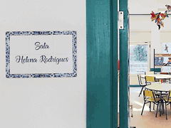 Placa de azulejos «Sala Helena Rodrigues» ao lado da entrada para sala com mesas redondas, estantes, janelas e borboletas coloridas pensuradas no teto