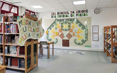 Estantes com livros (uma com cartaz grande) e, ao fundo, parede com grande painel com árvore formada por quadrados