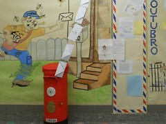 Marco de correio vermelho com cartas penduradas até ao teto; atrás, painéis com carteiro e várias cartas afixadas