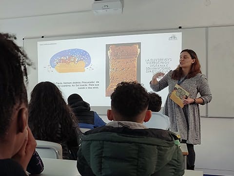 Senhora em frente a alunos sentados com livro numa mão junto a quadro branco onde estão projetados uma ilustração e uam pedra inscrita