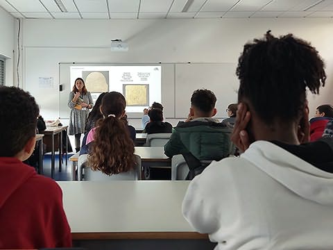 Alunos sentados numa sala de aula em frente a senhora com livro e com projeção de imagens atrás