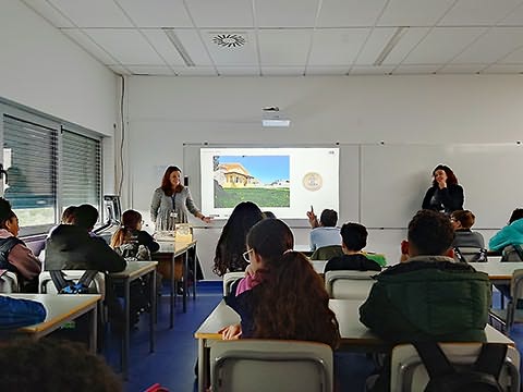 Duas senhoras junto a quadro de sala de aula, um aluno à frente de dedo no ar e outros sentados