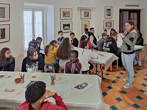 Muitos alunos sentados em mesas dispostas em arco com panos, ilustrações, lápis de cor..