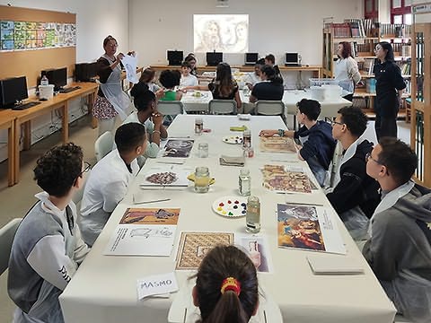 Numa biblioteca, muitos alunos sentados em mesas grandes com ilustrações coloridas, tintas e frascos; senhora mostrando folha; imagem projetada na parede ao fundo