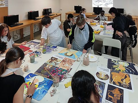 Vista de cima de duas mesas grandes cheias de pinturas e ilustrações, com alguns alunos sentados e outros em pé