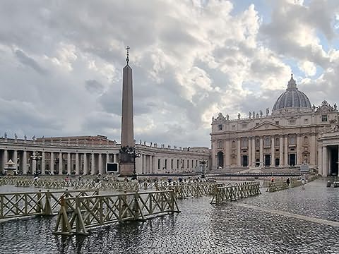 Grande praça de chão molhado com obelisco a meio e à volta edifícios com muitas colunas