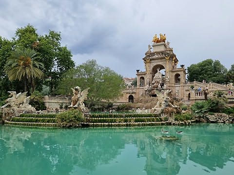 Estrutura com arcos e est&aacute;tua dourada no topo, criaturas aladas ao lado, escadaria e lago verde-azulado em primeiro plano com arvoredo em fundo