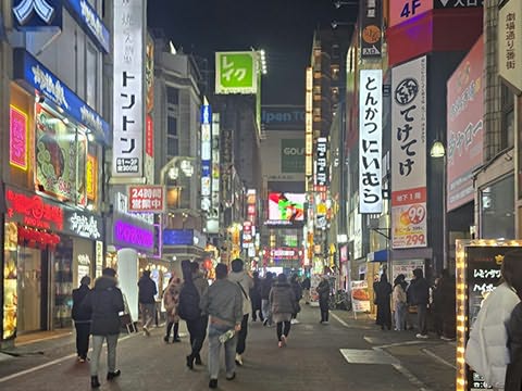 Rua comercial iluminada &agrave; noite, com v&aacute;rias pessoas a caminhar entre lojas e letreiros luminosos