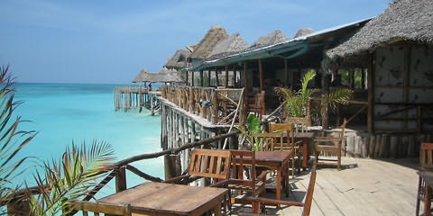 Mesas e cadeiras de madeira à frente de bangalós de telhados de palha cinzenta em espécie de cais sobre estacas em mar turquesa claro