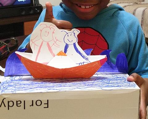 Caixa de sapatos com tampa colorida a azul, com barco de papel colorido a laranja, 2 pescadores desenhados a marcador e recortados e ondas coloridas a azul