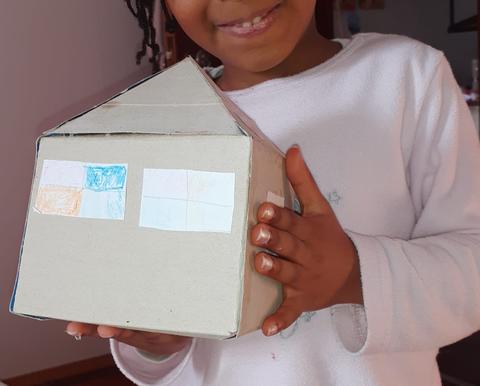 Caixa de cartão recortada em forma de casa (tridimensional) com janelas (desenhadas em papel a lápis de cor) recortadas e coladas