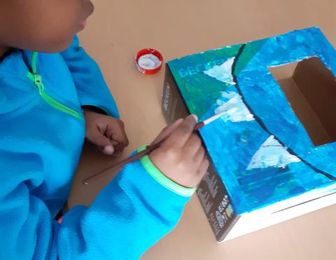 Menino pinta caixa de cartão recortada com guache azul, verde e branco