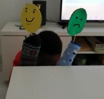 Menino meio escondido atrás de móvel com 2 fantoches feitos com meias e caras de cartão coloridas a marcadores verde e amarelo